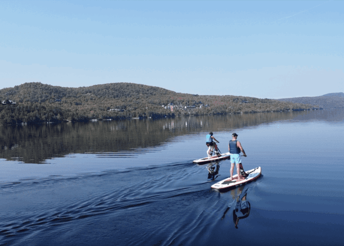 Two people paddleboard on a calm lake, leaving gentle ripples behind them, with tree-covered hills visible in the background under a clear blue sky.