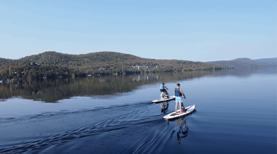 Deux personnes pratiquent des sports nautiques, font du paddleboard sur un lac calme entouré de collines couvertes d’arbres sous un ciel bleu clair. Deux personnes pratiquent des sports nautiques, font du paddleboard sur un lac calme entouré de collines couvertes d'arbres sous un ciel bleu clair.