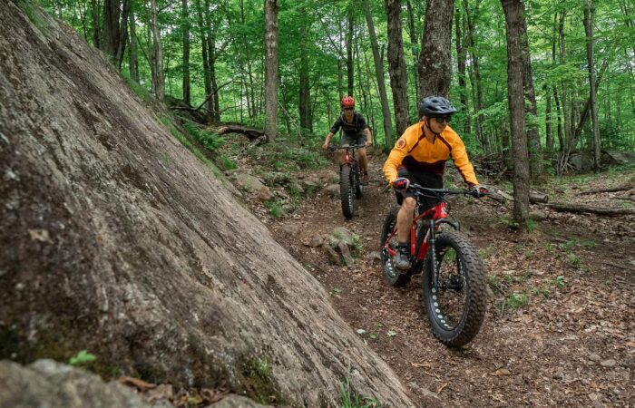 Two people ride fat-tire mountain bikes on a dirt trail through a dense, green forest with rocks and trees around them.