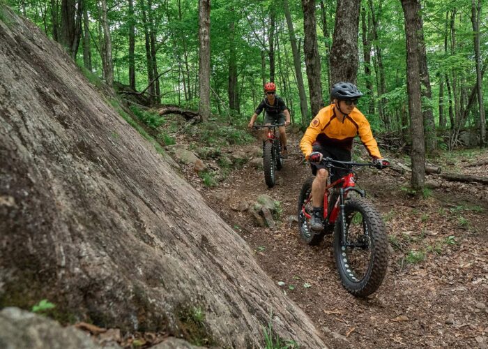 Two people ride fat-tire mountain bikes on a dirt trail through a dense, green forest with rocks and trees around them.
