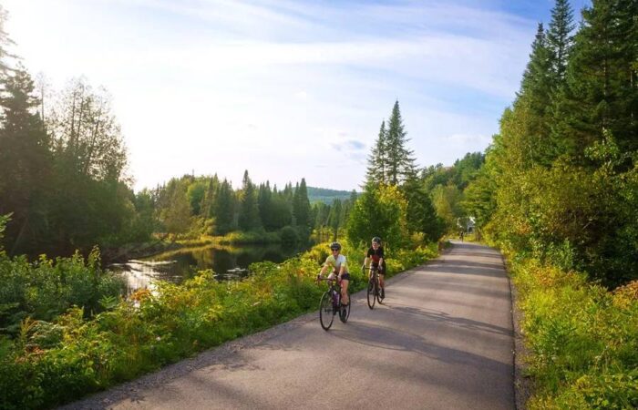 Deux personnes font de l'excursion à vélo sur un chemin pavé entouré d'arbres et de verdure, avec un petit plan d'eau sur la gauche et un ciel dégagé au-dessus.