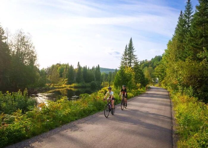 Two people ride bicycles on a paved path surrounded by trees and greenery, with a small body of water on the left and a clear sky above.