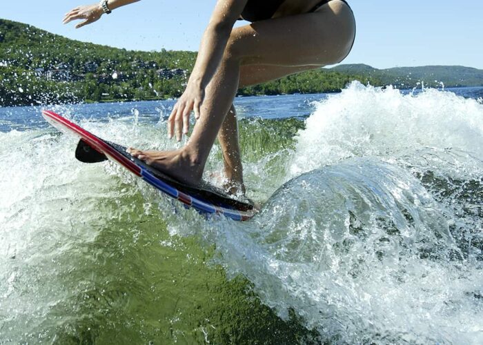 A person in a black swimsuit rides a wave on a wakesurf board on a lake, surrounded by hills under a clear blue sky.