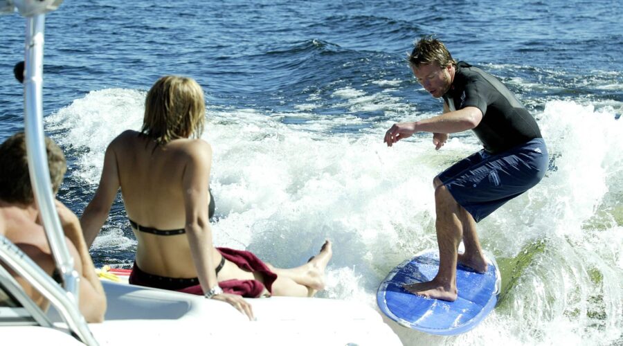 A man surfs on a blue board behind a boat while a woman in a bikini and another person watch from the boat on a sunny day. A man surfs on a blue board behind a boat while a woman in a bikini and another person watch from the boat on a sunny day.