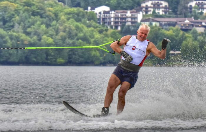Un homme d'un certain âge s'adonnant au ski nautique sur un lac, tenant une corde de remorquage, avec des arbres et des bâtiments visibles à l'arrière-plan - une journée parfaite pour les sports nautiques.
