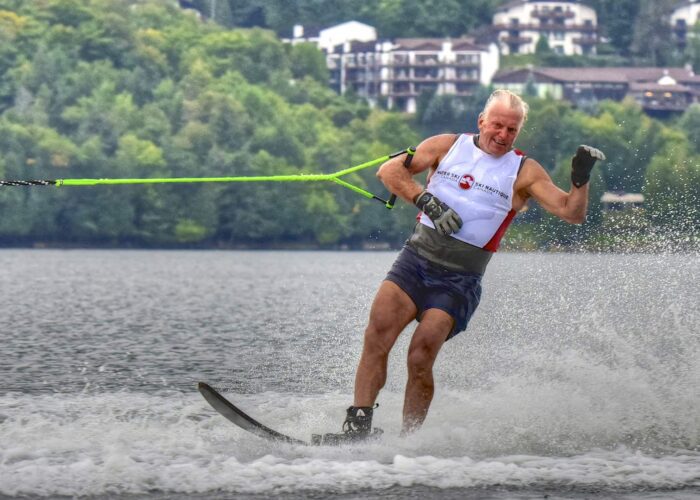 An older man wearing a life vest water skis on a lake, holding a tow rope, with splashes around him and buildings visible in the background.