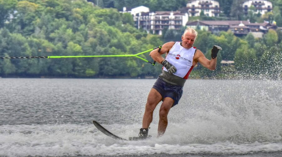 Un homme d'un certain âge s'adonnant au ski nautique sur un lac, tenant une corde de remorquage, avec des arbres et des bâtiments visibles à l'arrière-plan - une journée parfaite pour les sports nautiques.