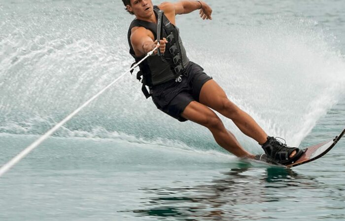 A person wearing a life vest is waterskiing on a lake, leaning to one side while holding onto a tow rope, with trees visible in the background.