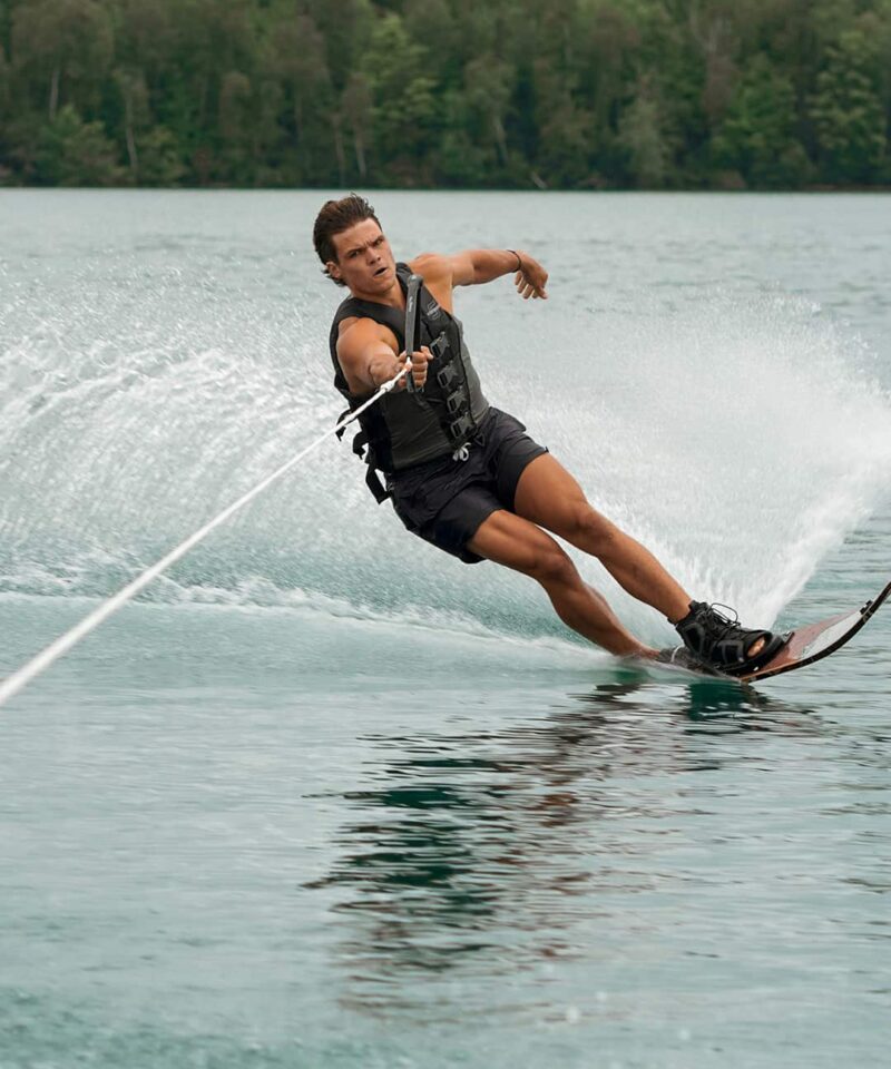 A person wearing a life vest is waterskiing on a lake, leaning to one side while holding onto a tow rope, with trees visible in the background.