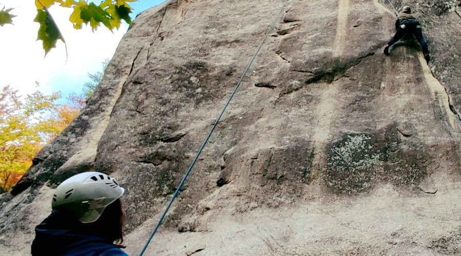 A person climbs a steep rock face while another person wearing a helmet belays from below; trees and blue sky are visible at the top.