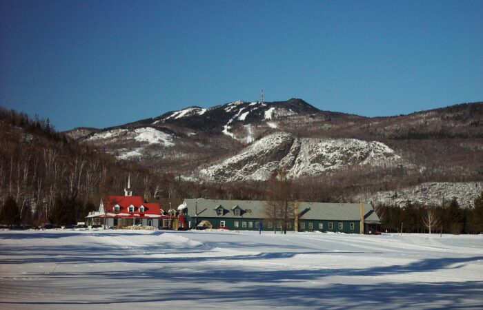Snow-covered buildings sit in front of a mountain with ski trails and a communication tower under a clear blue sky at Domaine Saint-Bernard Park. Snow-covered buildings sit in front of a mountain with ski trails and a communication tower under a clear blue sky at Domaine Saint-Bernard Park.