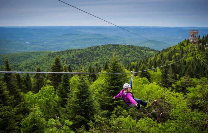 A person wearing a helmet and harness zip-lines over a lush green forest with mountains and a cloudy sky in the background. A person wearing a helmet and harness zip-lines over a lush green forest with mountains and a cloudy sky in the background.