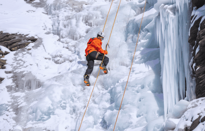 A person wearing an orange jacket and helmet climbs an icy, snow-covered slope using ropes and climbing gear.