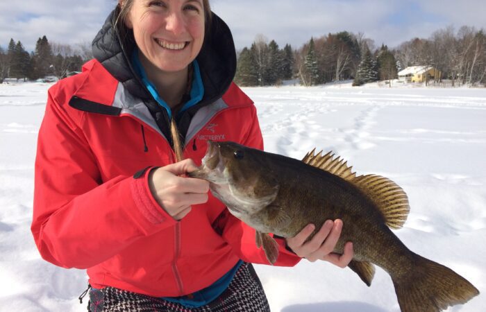 A woman in a red jacket holds a large fish while sitting on snow, with trees and houses in the background.