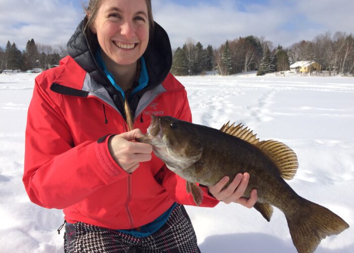 A woman in a red jacket holds a large fish while sitting on snow, with trees and houses in the background.