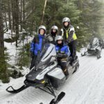 Four people wearing helmets and winter gear sit on two snowmobiles parked on a snowy forest trail, ready for snowmobiling tours with all the necessary equipment, surrounded by trees and fresh snow. Four people wearing helmets and winter gear sit on two snowmobiles parked on a snowy forest trail, ready for snowmobiling tours with all the necessary equipment, surrounded by trees and fresh snow.