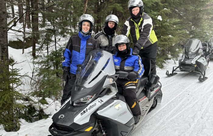 Four people wearing helmets and winter gear sit on two snowmobiles parked on a snowy forest trail, ready for snowmobiling tours with all the necessary equipment, surrounded by trees and fresh snow.
