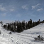 Three people ride snowmobiles along scenic snowmobile trails surrounded by pine trees and mounds of snow under a blue sky. Three people ride snowmobiles along scenic snowmobile trails surrounded by pine trees and mounds of snow under a blue sky.