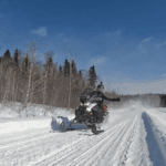 A person rides a snowmobile on a snowy trail through a forest with clear blue sky overhead, enjoying the best time for snowmobiling during the vibrant Quebec snowmobile season. A person rides a snowmobile on a snowy trail through a forest with clear blue sky overhead, enjoying the best time for snowmobiling during the vibrant Quebec snowmobile season.