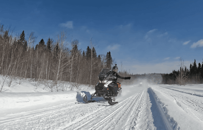 A person rides a snowmobile on a snowy trail through a forest with clear blue sky overhead, enjoying the best time for snowmobiling during the vibrant Quebec snowmobile season.