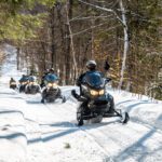 A group of people enjoy a beginner snowmobile tour through a snowy forest trail, with trees on both sides and sunlight filtering through the branches. A group of people enjoy a beginner snowmobile tour through a snowy forest trail, with trees on both sides and sunlight filtering through the branches.
