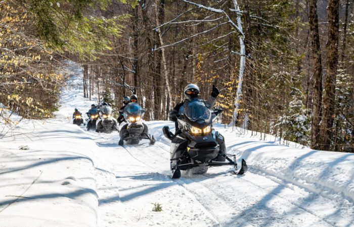 A group of people enjoy a beginner snowmobile tour through a snowy forest trail, with trees on both sides and sunlight filtering through the branches.