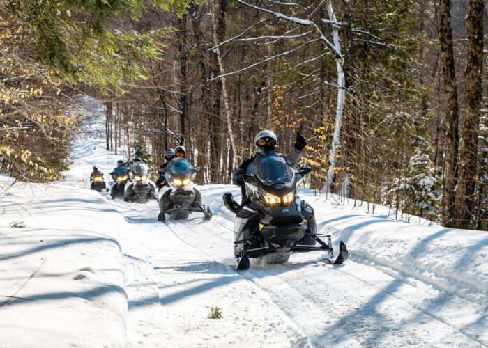 A group of people enjoy a beginner snowmobile tour through a snowy forest trail, with trees on both sides and sunlight filtering through the branches.