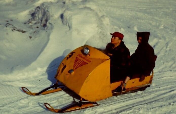 Two people ride a vintage yellow snowmobile across a snowy landscape, celebrating the rich history of snowmobiling in Canada.