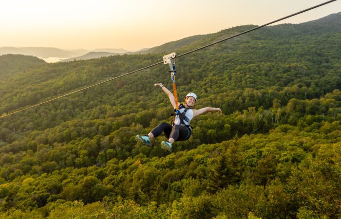 A person wearing a helmet and harness rides a zip line above a dense green forest with hills and a sunset in the background. A person wearing a helmet and harness rides a zip line above a dense green forest with hills and a sunset in the background.