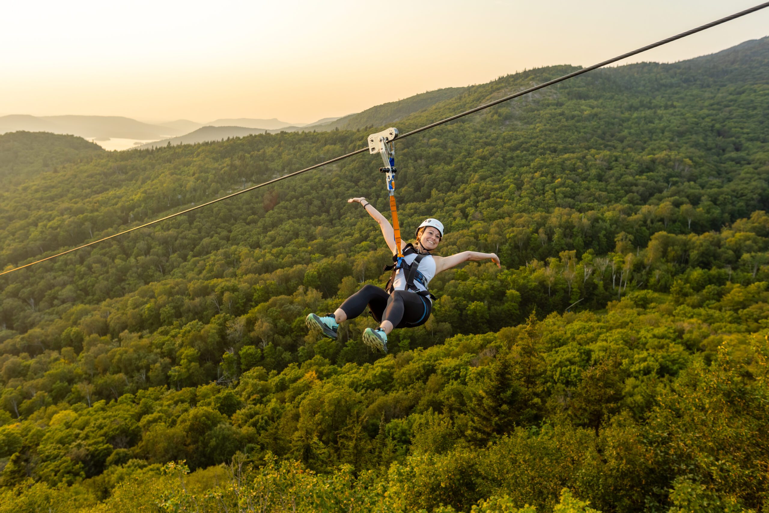A person wearing a helmet and harness rides a zip line above a dense green forest with hills and a sunset in the background. A person wearing a helmet and harness rides a zip line above a dense green forest with hills and a sunset in the background.