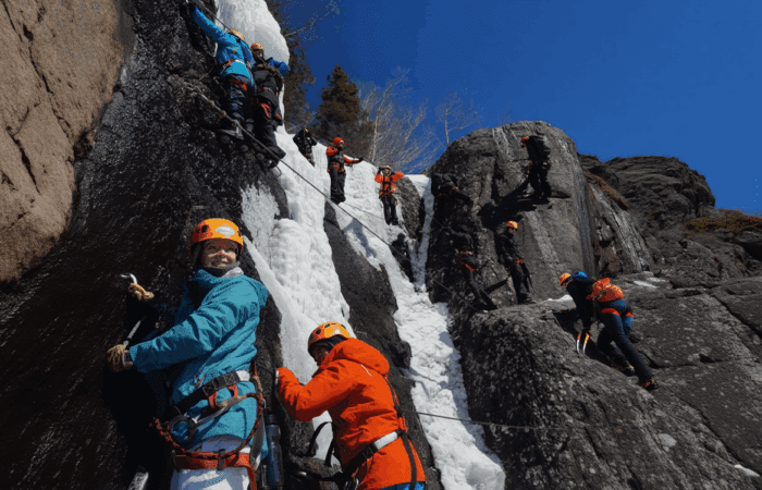 A group of people in helmets and climbing gear scale a rocky, icy cliff face under a clear blue sky.