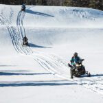 Three people ride snowmobiles on a snowy hill, following parallel tracks under a clear sky with trees in the background—reminding riders to check snowmobile driving requirements before heading out. Three people ride snowmobiles on a snowy hill, following parallel tracks under a clear sky with trees in the background—reminding riders to check snowmobile driving requirements before heading out.