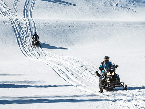 Three people ride snowmobiles on a snowy hill, following parallel tracks under a clear sky with trees in the background—reminding riders to check snowmobile driving requirements before heading out.