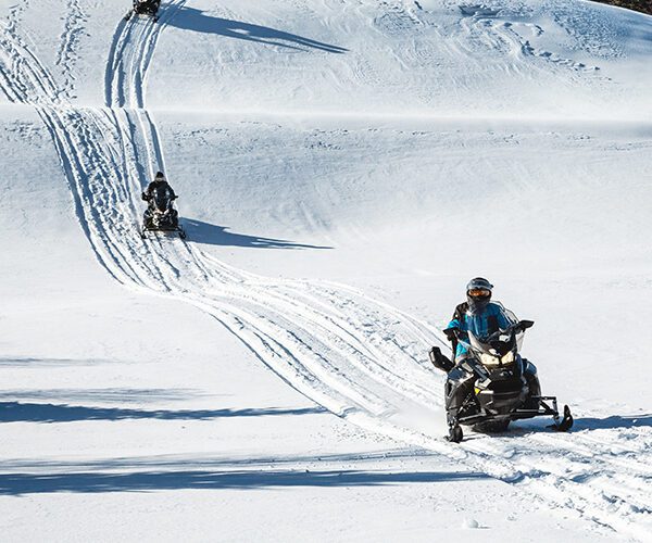 Three people ride snowmobiles on a snowy hill, following parallel tracks under a clear sky with trees in the background—reminding riders to check snowmobile driving requirements before heading out.