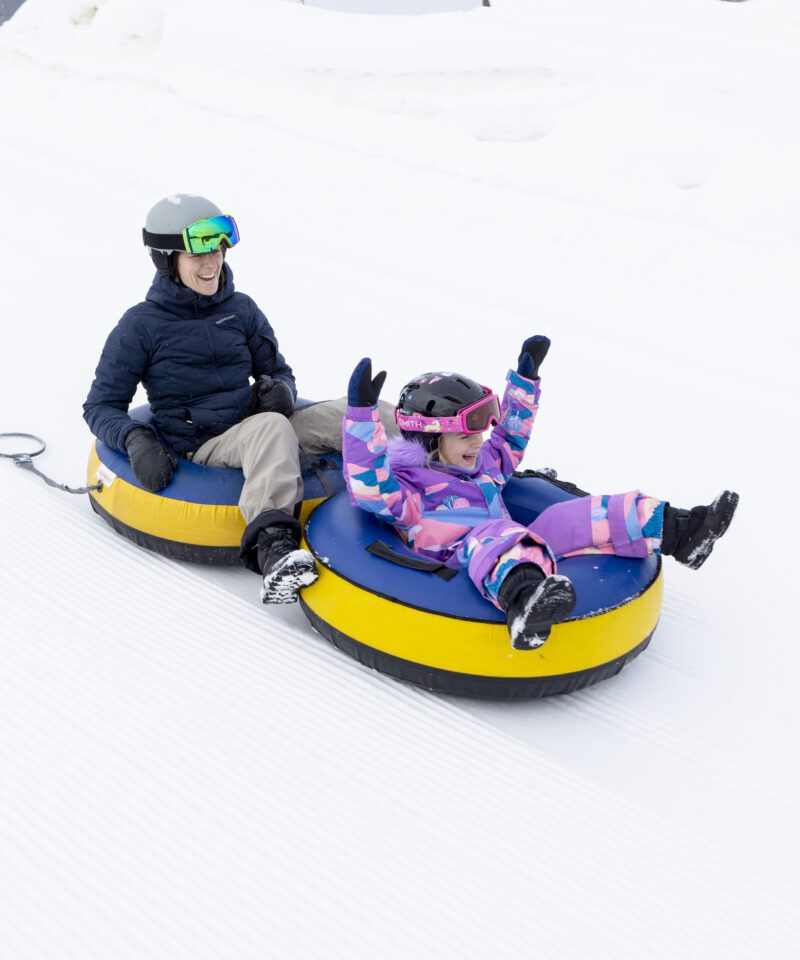 An adult and a child, both wearing helmets and winter clothing, ride yellow snow tubes down a snowy hill.