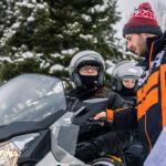 A man in an orange and black jacket is instructing two people on a snowmobile, discussing the snowmobile minimum age. All are wearing helmets and winter gear, with snowy trees in the background. A man in an orange and black jacket is instructing two people on a snowmobile, discussing the snowmobile minimum age. All are wearing helmets and winter gear, with snowy trees in the background.