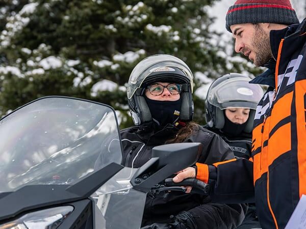A man in an orange and black jacket is instructing two people on a snowmobile, discussing the snowmobile minimum age. All are wearing helmets and winter gear, with snowy trees in the background.
