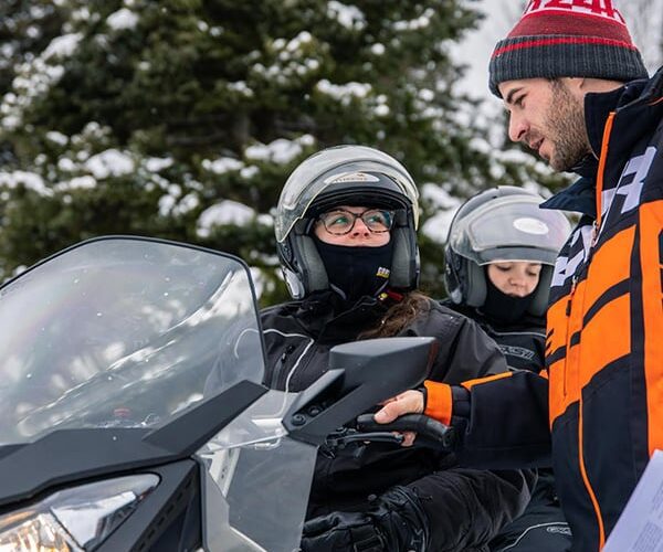 A man in an orange and black jacket is instructing two people on a snowmobile, discussing the snowmobile minimum age. All are wearing helmets and winter gear, with snowy trees in the background.