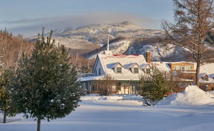 Maison et arbres enneigés dans un paysage hivernal près de Mont-Tremblant, avec les montagnes en arrière-plan sous un ciel clair - parfait pour une paisible randonnée en raquettes.