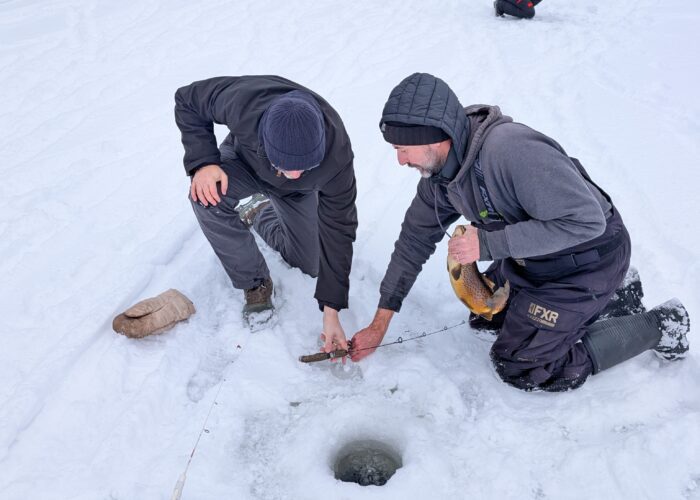 Two people kneel on a snowy mountain lake, pulling a fish from an ice fishing hole. Fishing gear and a thermos are nearby; other people and tents dot the frozen landscape in the background.