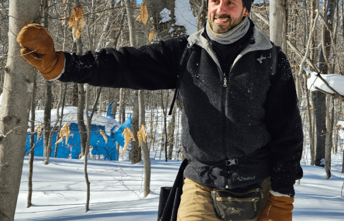 A man wearing winter clothing and mittens stands in a snowy forest, touching a tree with one hand. Blue structure is visible in the background.