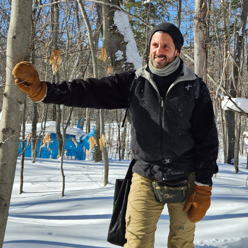 A man wearing winter clothing and mittens stands in a snowy forest, touching a tree with one hand. Blue structure is visible in the background. A man wearing winter clothing and mittens stands in a snowy forest, touching a tree with one hand. Blue structure is visible in the background.