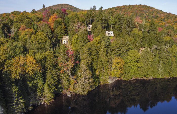 Vue aérienne de trois petites cabanes dans les arbres nichées au milieu d'arbres denses, verts et automnaux, sur une colline au-dessus d'un lac calme, avec des montagnes en arrière-plan.