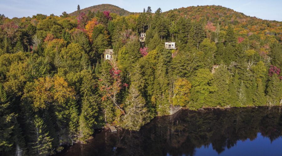 Houses are partially visible among dense, colorful autumn trees on a hillside above a dark, calm lake. Houses are partially visible among dense, colorful autumn trees on a hillside above a dark, calm lake.