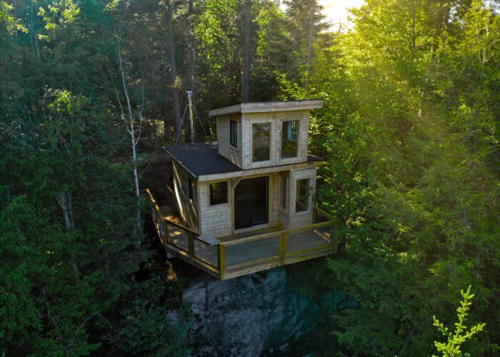 A small two-story wooden cabin with large windows and a deck is nestled among dense green trees on a rocky hillside.