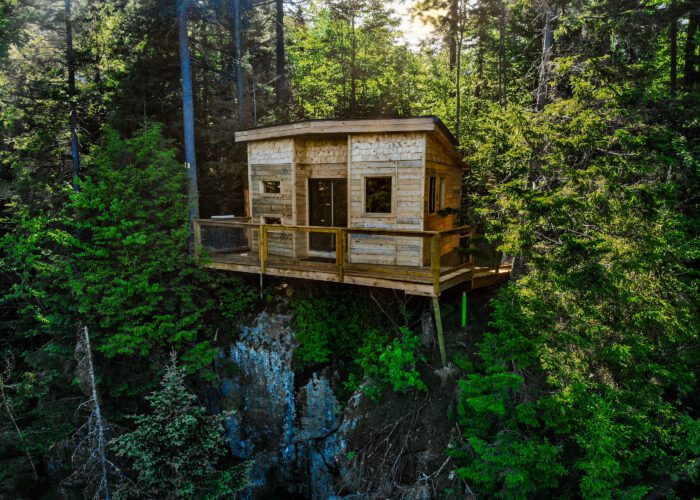 A modern wooden cabin built on stilts stands on the edge of a rocky cliff, surrounded by dense, green forest.