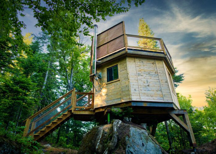 A small wooden cabin with a rooftop deck stands elevated on rocks among trees, with a staircase leading up to it under a partly cloudy sky.