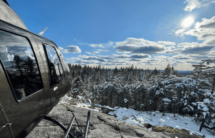 A Summit Flight black helicopter is parked on a rocky ledge overlooking a snowy forest under a partly cloudy sky with the sun shining.