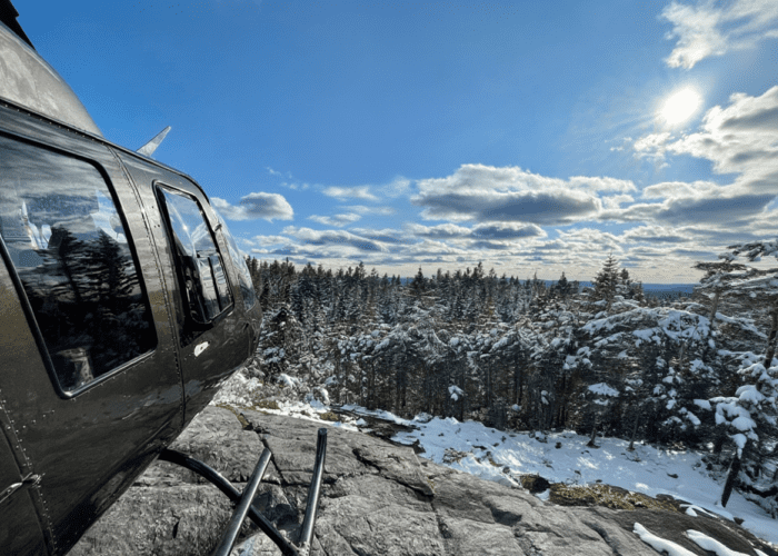 A Summit Flight black helicopter is parked on a rocky ledge overlooking a snowy forest under a partly cloudy sky with the sun shining.