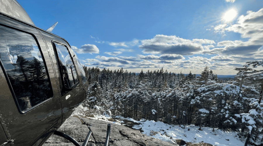 Un Hélicoptère noir est garé sur une surface rocheuse, surplombant une forêt enneigée sous un ciel pratiquement dégagé et un soleil radieux - parfait pour un vol au sommet.
